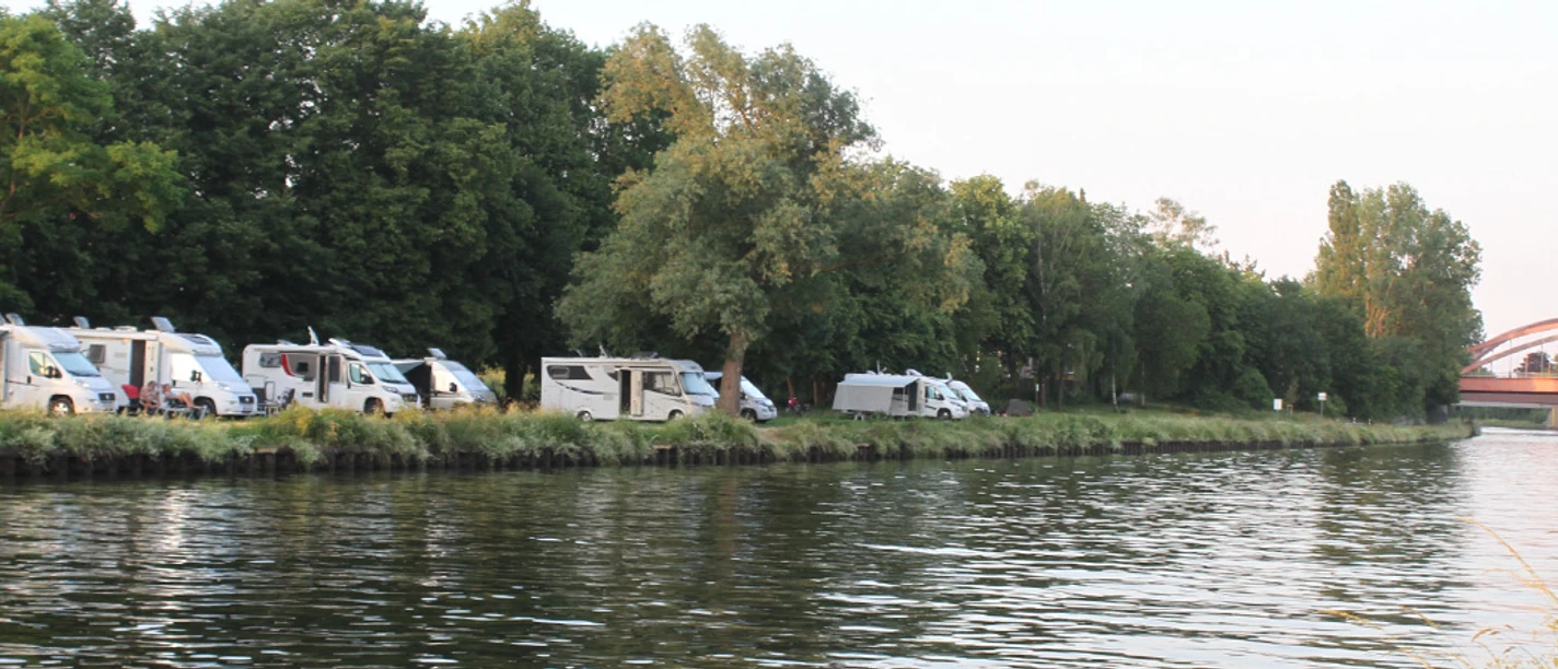 Wohnmobile02.jpg Wohnmobile parken entlang eines Flusses, von Bäumen gesäumt; im Hintergrund eine Brücke sichtbar.