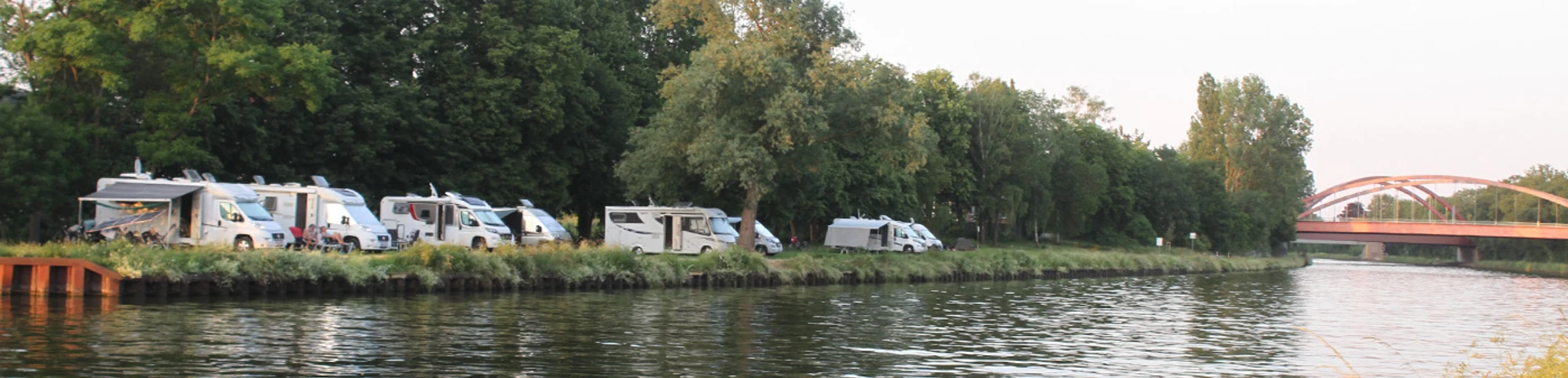 Wohnmobile02.jpg Wohnmobile parken entlang eines Flusses, von Bäumen gesäumt; im Hintergrund eine Brücke sichtbar.