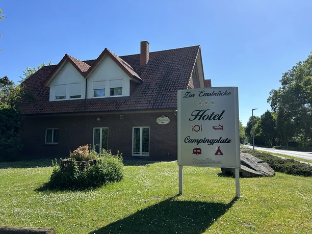 Campingplatz Zur Emsbrücke ©Emsland Tourismus GmbH (5).jpeg Backsteinhaus mit rotem Dach und Schild des Campingplatzes Zur Emsbrücke bei sonnigem Wetter