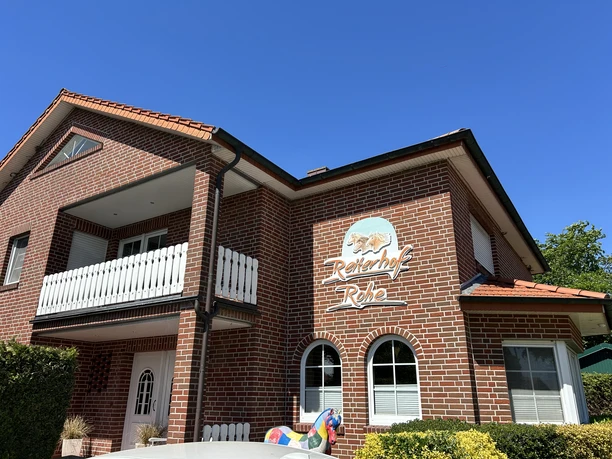 Reiterhof Rohe ©Emsland Tourismus GmbH Backsteingebäude mit Balkon und weißem Zaun, Schriftzug Reiterhof Rohe an der Fassade, blauer Himmel