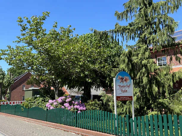 Reiterhof Rohe ©Emsland Tourismus GmbH Eingang des Reiterhofs Rohe mit grünem Zaun, Blütensträuchern und Hinweisschild bei Sonnenschein.