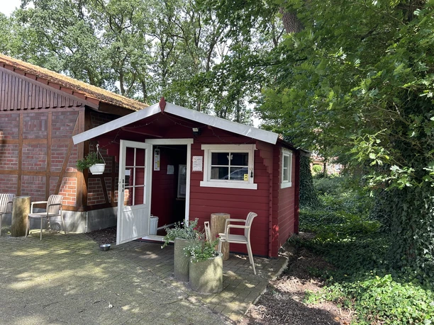Kleines rotes Holzhaus mit offener Tür, Sitzgelegenheiten und Blumenschmuck unter Bäumen.