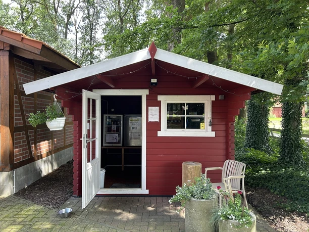 Rotes Holzhaus mit offener Tür, Pflanzen vor dem Eingang und Bäumen im Hintergrund.
