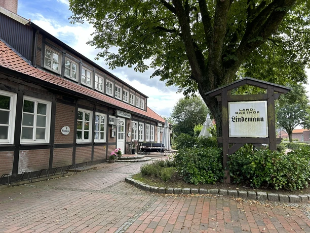 Hof Lindemann Heimatverein Gersten ©Emsland Tourismus GmbH (3).JPEG Backsteinfachwerkhaus mit roten Dachziegeln, großem Baum und Schild Landgasthof Lindemann an Weg.
