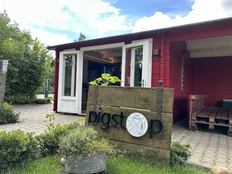 Hof Pleus-Pigstop, Bawinkel ©Emsland Tourismus GmbH (3).JPEG Rotes Holzgebäude mit offener Veranda, Sitzbank aus Paletten und Holzschild mit Aufschrift Pigstop.