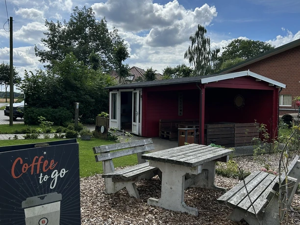 Hof Pleus-Pigstop, Bawinkel ©Emsland Tourismus GmbH (6).JPEG Holztische mit Bänken vor rotem Hofgebäude, daneben Schild mit Aufschrift Coffee to go.