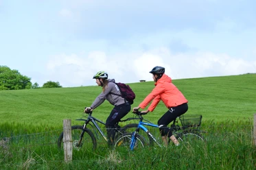 Raderlebnis Schieder-Schwalenberg Zwei Radfahrer mit Helmen genießen eine Fahrt auf einem grünen Weg bei bewölktem Himmel.