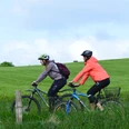 Raderlebnis Schieder-Schwalenberg Zwei Radfahrer mit Helmen genießen eine Fahrt auf einem grünen Weg bei bewölktem Himmel.