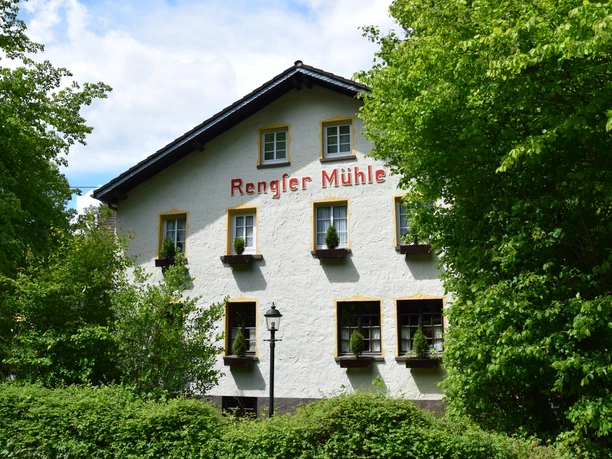 Rengser Mühle Rengser Mühle in Gummersbach, weißes Fachwerkhaus umgeben von üppigem Grün, blauer Himmel.