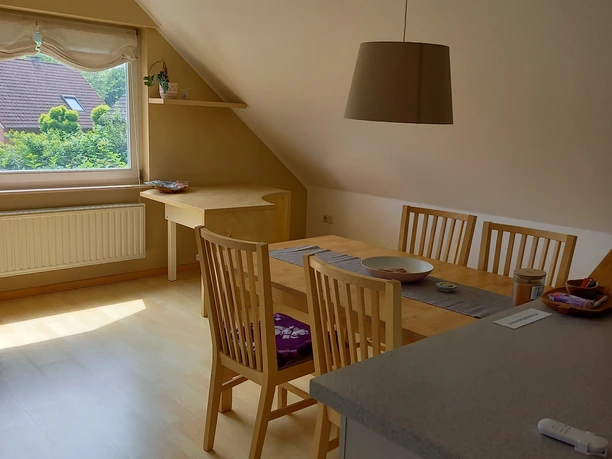 Helles, gemütliches Dachgeschosszimmer mit Esstisch, Stühlen, Fensterblick auf grüne Landschaft.