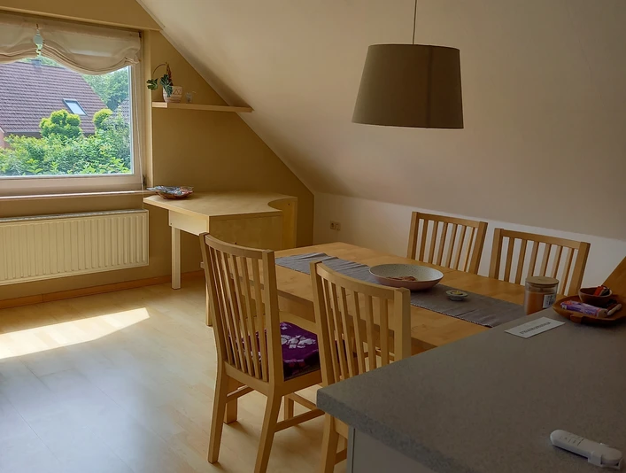 Ferienwohnung Schwarz.jpg Helles, gemütliches Dachgeschosszimmer mit Esstisch, Stühlen, Fensterblick auf grüne Landschaft.
