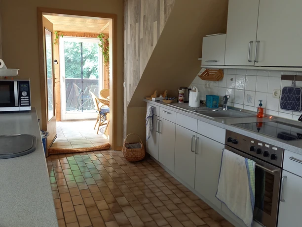 Ferienwohnung Schwarz Küche.jpg Helle Küche mit L-förmiger Arbeitsplatte, weißen Schränken, Fensterausblick auf grünen Balkon.