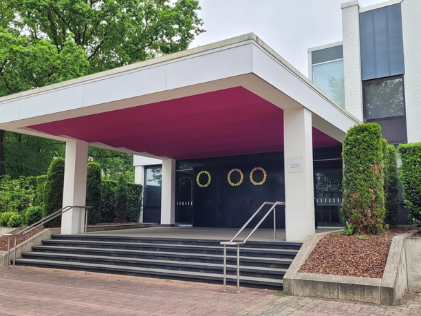 Der Eingang des Neuen Theaters Espelkamp mit modernen Linien und umgeben von gepflegter Bepflanzung.