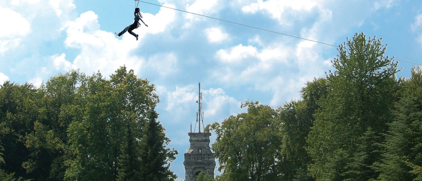 Seilbahn mit Bismarckturm im Waldkletterpark Velbert-Langenberg.jpg Eine Person schwebt mit einer Seilbahn über Baumwipfel vor dem Bismarckturm im Kletterpark.