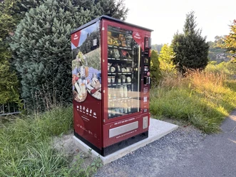 Proviantomat Standort in Pötzscha/Stadt Wehlen Ein roter Verkaufsautomat steht auf einem Betonfundament in einer grünen, ländlichen Umgebung, gefüllt mit verschiedenen Lebensmitteln.