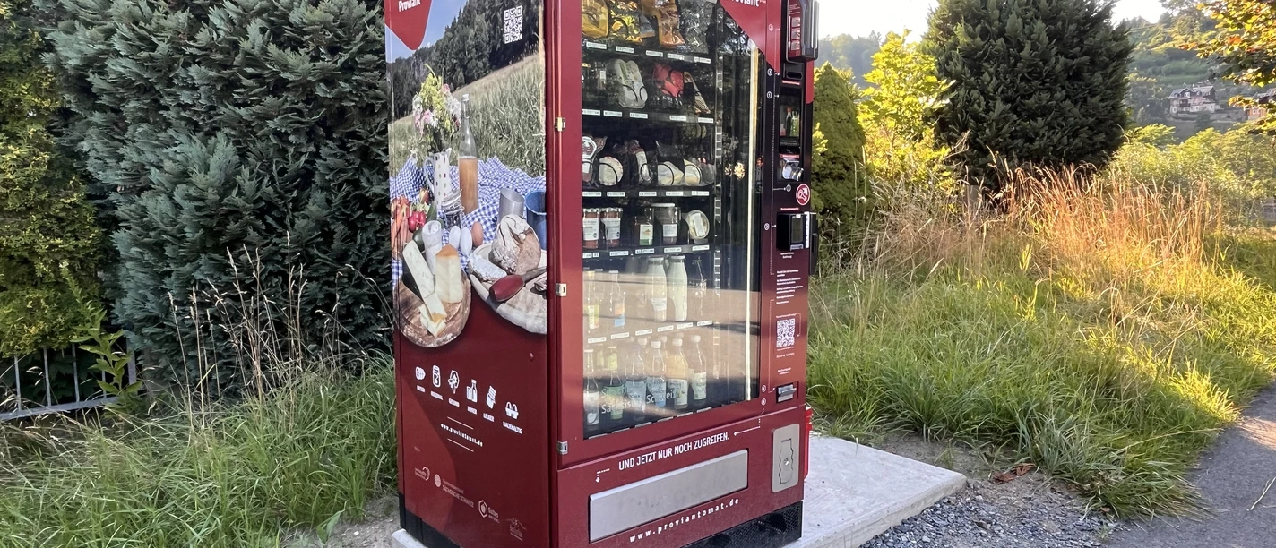 Proviantomat Standort in Pötzscha/Stadt Wehlen Ein roter Verkaufsautomat steht auf einem Betonfundament in einer grünen, ländlichen Umgebung, gefüllt mit verschiedenen Lebensmitteln.