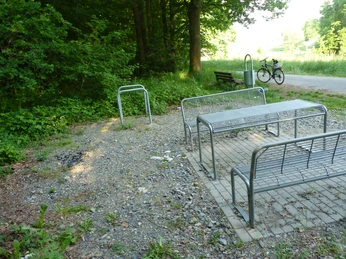 Zwei Metalltische und Bänke stehen auf gepflastertem Boden an einem Waldpfad mit Fahrrad.