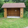 Hölzerner Unterstand mit überdachter Sitzgelegenheit, umgeben von grüner Vegetation am Wegesrand.