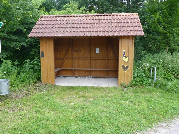 Hölzerner Unterstand mit überdachter Sitzgelegenheit, umgeben von grüner Vegetation am Wegesrand.