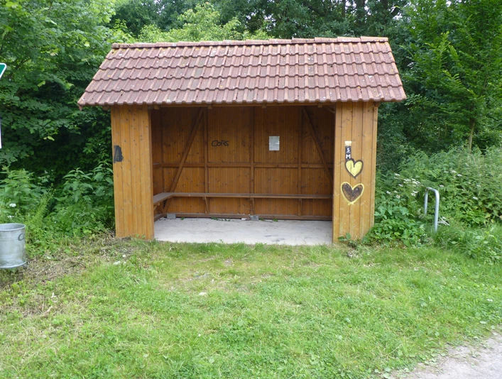 Hölzerner Unterstand mit überdachter Sitzgelegenheit, umgeben von grüner Vegetation am Wegesrand.