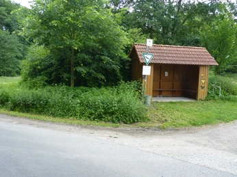 Bushaltestelle mit überdachtem Holzunterstand, umgeben von Bäumen und grüner Vegetation am Straßenrand.