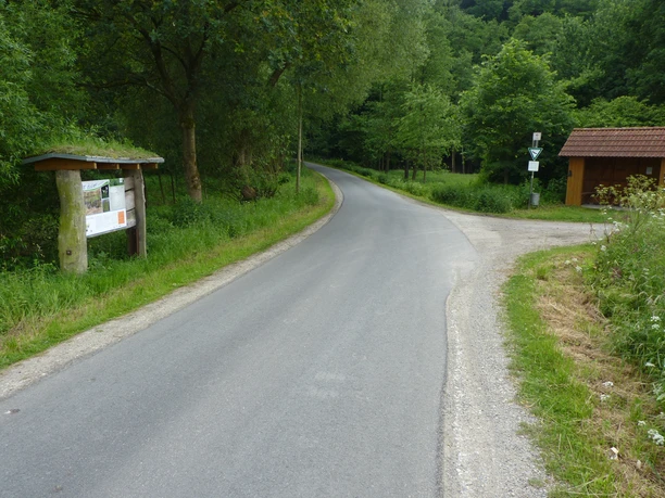 Eine asphaltierte Landstraße führt durch üppiges Grün, vorbei an einem Wegweiser und einem Holzunterstand.