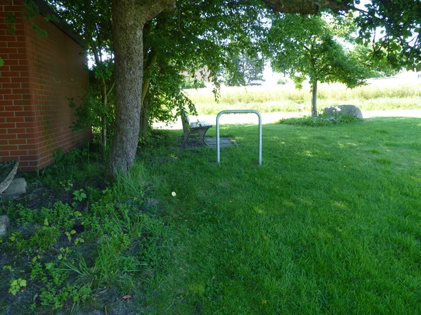 Unter einem Schattenbaum steht eine Holzbank neben einer Backsteinmauer, umgeben von grüner Wiese.