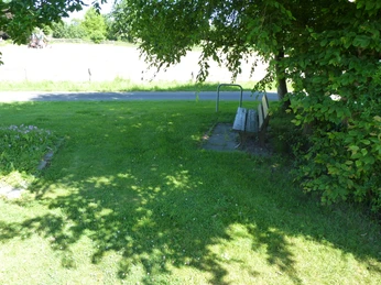 Parkbank unter großen Schatten spendenden Bäumen neben einer Wiese und vor einer ruhigen Landstraße.