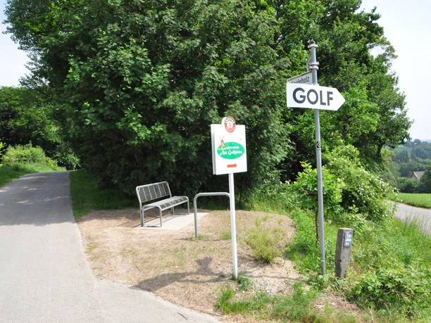 Wegweiser zum Golfplatz neben einer Parkbank an einem ländlichen Weg, umgeben von grünen Bäumen.