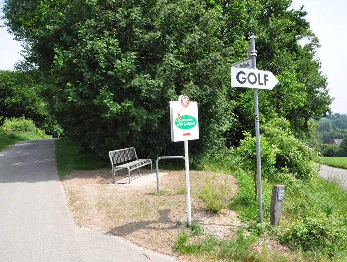 Wegweiser zum Golfplatz neben einer Parkbank an einem ländlichen Weg, umgeben von grünen Bäumen.