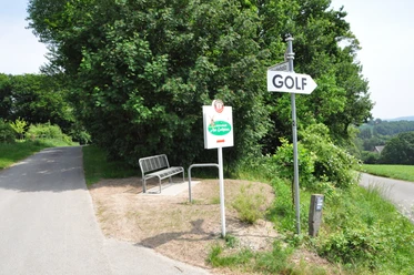 DSC_0062.JPG Wegweiser zum Golfplatz neben einer Parkbank an einem ländlichen Weg, umgeben von grünen Bäumen.