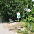 DSC_0062.JPG Wegweiser zum Golfplatz neben einer Parkbank an einem ländlichen Weg, umgeben von grünen Bäumen.
