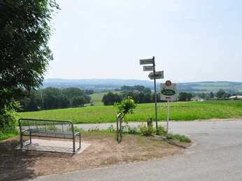 Eine Metallbank steht an einem Wegesrand mit Schildern, umgeben von grünen Feldern und Hügeln.