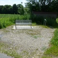 Eine Metallbank steht auf einem Schotterplatz am Rand eines Feldes, umgeben von grüner Vegetation.