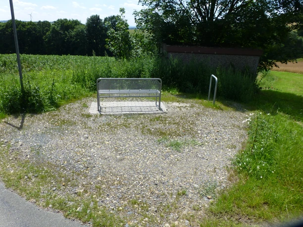 Eine Metallbank steht auf einem Schotterplatz am Rand eines Feldes, umgeben von grüner Vegetation.