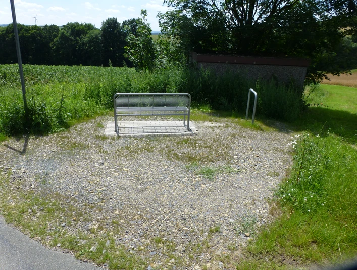 P1120720.JPG Eine Metallbank steht auf einem Schotterplatz am Rand eines Feldes, umgeben von grüner Vegetation.