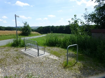 P1120730.JPG Metallbank mit Rückenlehne auf einem Schotterplatz, umgeben von grüner Landschaft und Feldern.