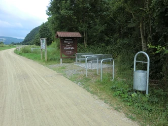Fahrradabstellplätze und Infotafel am Waldrand neben einem Schotterweg, Mülleimer rechts im Bild.