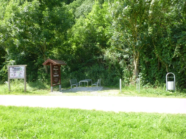 Rastplatz mit zwei Bänken und Tisch, umgeben von grünen Bäumen und Infotafeln auf einem Radweg.