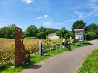 Radfahrerin an einem Rastplatz mit Infotafel und Fahrradständer neben einem Feld bei sonnigem Wetter.