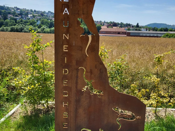 Metallschild mit ausgestanzten Eidechsen, auf grünem Feld vor einem klaren blauen Sommerhimmel.