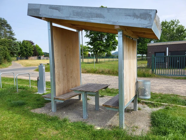 Ein einfacher Rastplatz mit Dach, Tisch und zwei Bänken auf einer Wiese, umgeben von Bäumen.