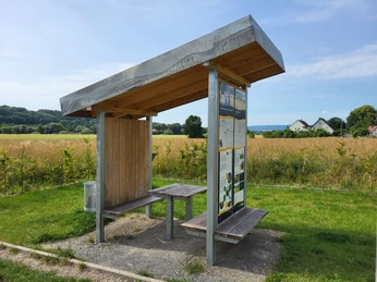 Holzüberdachte Sitzbank an einer ruhigen Feldwegkreuzung, flankiert von Infotafel und Mülleimer.