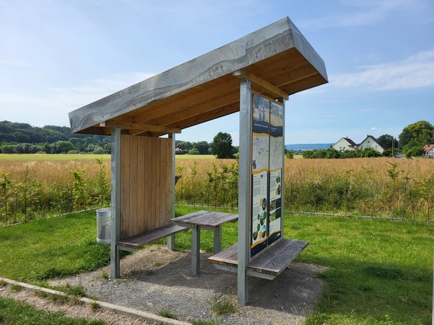 Holzüberdachte Sitzbank an einer ruhigen Feldwegkreuzung, flankiert von Infotafel und Mülleimer.