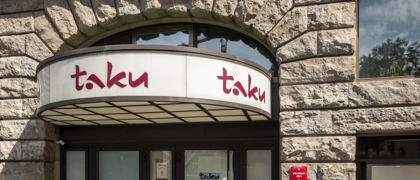 Eingang des eleganten Restaurants Taku mit Steinmauerwerk und einer modernen Glasfront.