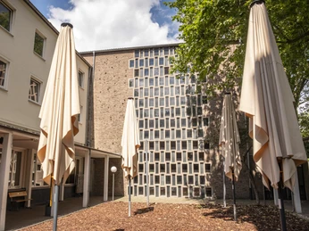 Luther Kirche Das Bild zeigt den Vorderhof der Luther Kirche mit markanten quadratischen Fensterstrukturen an der Fassade. Mehrere gefaltete Sonnenschirme stehen im gepflasterten und von Grün umgebenen Bereich.The picture shows the front courtyard of the Luther Church with striking square window structures on the façade. Several folded parasols stand in the paved area surrounded by greenery.
