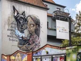 Cafe Friedolin BuezE Mosaikverzierte Mauer und im Hintergrund eine mit kunstvollem Graffiti versehene Hauswand.Mosaic-decorated wall and, in the background, a house wall with artistic graffiti.