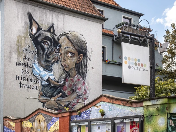 Cafe Friedolin BuezE Mosaikverzierte Mauer und im Hintergrund eine mit kunstvollem Graffiti versehene Hauswand.