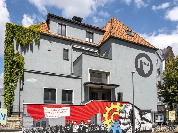 Cafe Friedolin BuezE Modernes graues Gebäude mit dem Logo des Bürgerzentrums Ehrenfeld. Im Vordergrund eine Mauer mit sozialkritischem Graffiti.