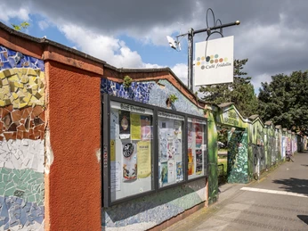 Cafe Fridolin Buetze Kunstvoll verzierte Mauer mit kleinen Mosaiksteinen mit Zugang zum Park. Ein Schaukasten mit Infoblättern.Artfully decorated wall with small mosaic stones, with access to the park. A display case with information sheets.